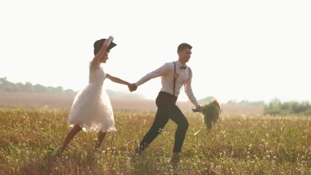 The newlyweds are running in the sunny field. The bride is throwing the black hat while the groom is holding the wedding bouquet. — Stock Video