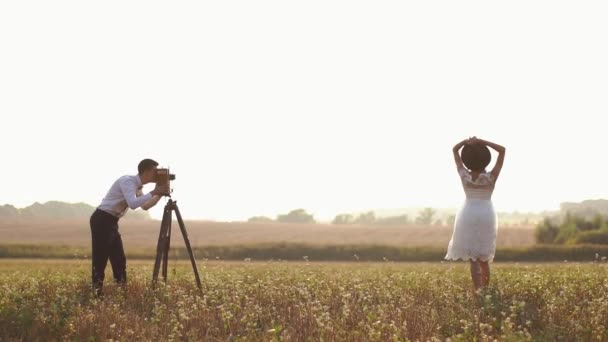 La vista lateral del novio tomando fotos de la novia posando usando la vieja cámara vintage en el campo soleado . — Vídeo de stock