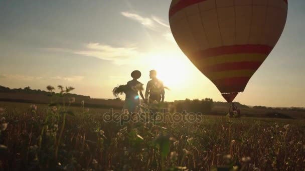 A romantikus elülső kilátás nyílik a wintage öltözött ifjú kezét, és séta a mező alatt a naplemente, a háttérben a színes hőlégballon. — Stock videók