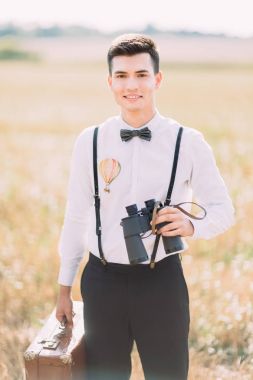 Dürbün ve vintage çanta tutarak ve kameraya gülümseyen damat dikey yakın çekim portresi.