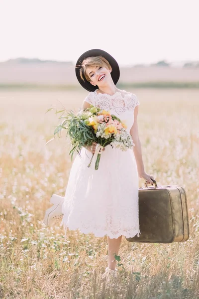 A noiva vestida vintage sorridente com o buquê de casamento e mala está posando para a câmera. A composição do campo ensolarado . — Fotografia de Stock