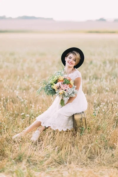 O retrato vertical da noiva sorridente com o buquê colorido sentado na mala vintage no campo de primavera . — Fotografia de Stock