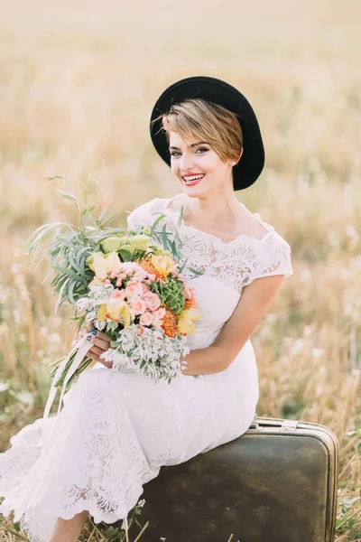La bella sposa seduta sulla valigia d'epoca, regge il bouquet colorato del matrimonio e sorride alla macchina fotografica. Il ritratto ravvicinato . — Foto Stock
