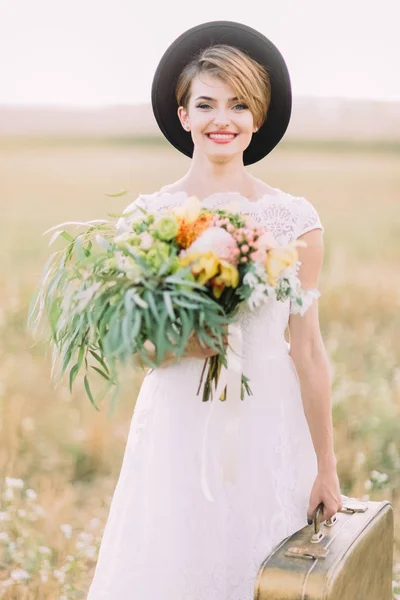 Il ritratto ravvicinato della bella donna sorridente in abito bianco che tiene il bouquet colorato e porta la valigia vintage sullo sfondo del campo soleggiato . — Foto Stock