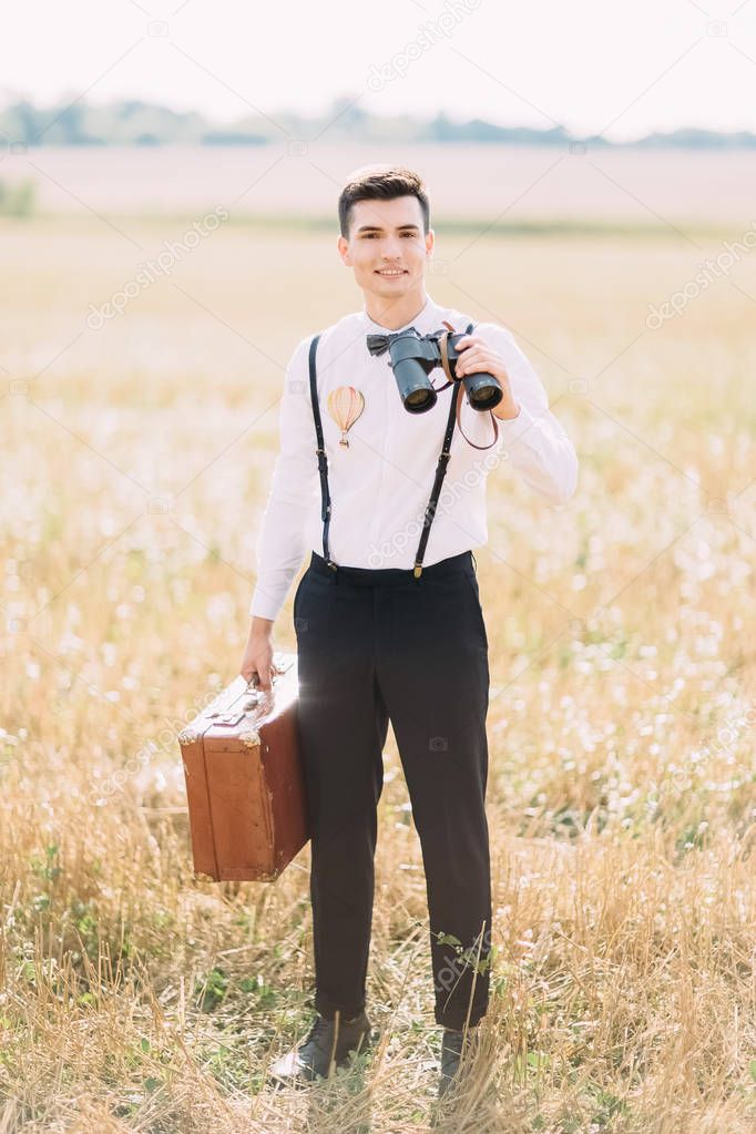 The smiling vintage dressed groom carrying the retro suitcase and binoculars is looking at the camera. The sunny field composition.