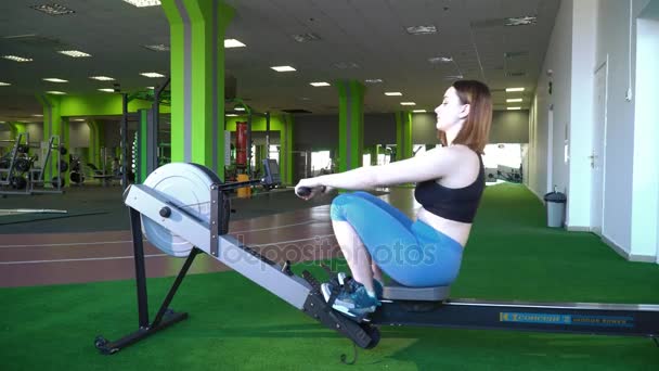 女人在蓝色的裤子和黑色上衣上划船机在绿色的健身房锻炼的侧视图. — 图库视频影像