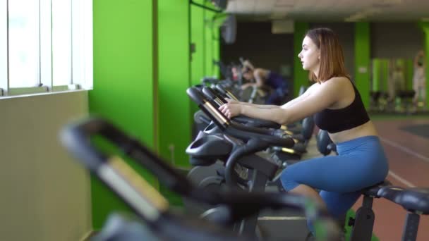 Το πορτρέτο οριζόντια πλευρά της γυναίκας στη μπλε παντελόνι και μαύρη κορυφή που εργάζονται για το ποδήλατο στο πράσινο γυμναστήριο. — Αρχείο Βίντεο