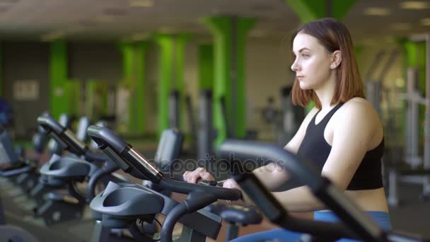 Het portret van de close-up zijde van de vrouw toning haar spieren op de hometrainer in de groene sportschool. — Stockvideo