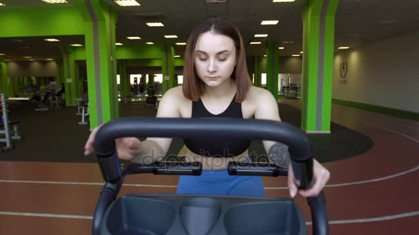 Närbild främre porträtt av kvinnan med blå ögon i blå byxor och svart topp uppvärmning på motionscykel i gröna gym. — Stockvideo