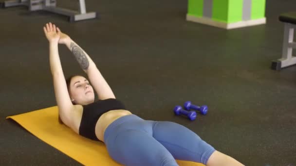 La joven está acostada en la colchoneta haciendo abdominales levantando las manos y las piernas en el gimnasio . — Vídeos de Stock