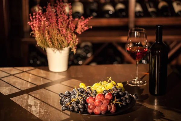 Composizione di bottiglia di vino, frutta di vetro e vaso di fiori posto su un tavolo di legno in una volta di vino . — Foto Stock