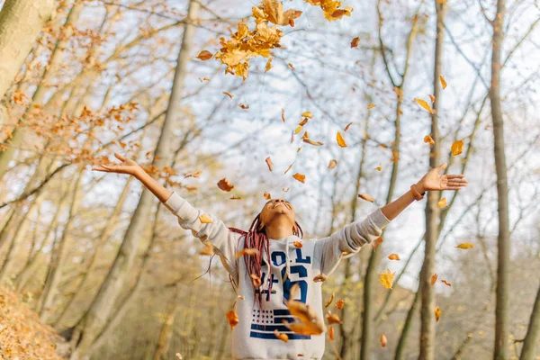 Atraente menina americana bonita africana em roupas esportivas brincando com um monte de folhas, desfrutando de dia quente de outono no parque. Divirta-se no outono — Fotografia de Stock