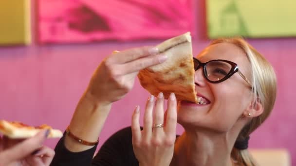 Close up shooting of beautiful woman. Pretty girl in glasses is sitting in a restaurant with colorful walls and eating pizza. She enjoys eating good food. — Stock Video