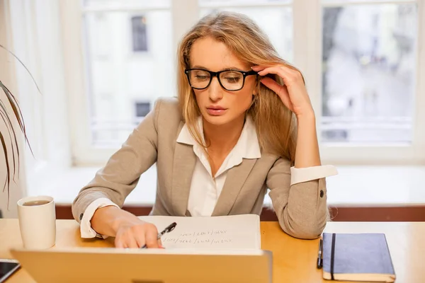 Atrativo jovem loiro de cabelos maravilhosos vestindo óculos e trabalhando com laptop. Mulher sentada em um escritório acolhedor agradável . — Fotografia de Stock