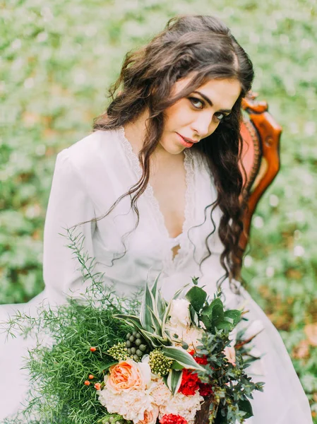 Härlig närbild porträtt av kvinnan i vit klänning, sitter på stolen, holding vackra buketten av blommor och tittar på kameran. Skogsnära läge. — Stockfoto