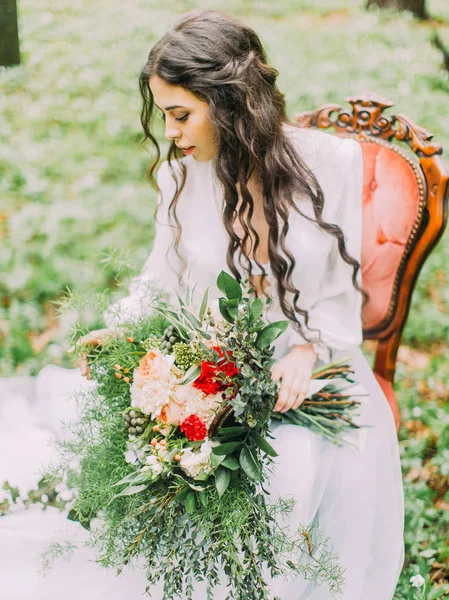El retrato lateral de la mujer con el pelo rizado largo en el vestido de novia blanco, sosteniendo el ramo de colores y sentado en la silla en el bosque verde de primavera . — Foto de Stock