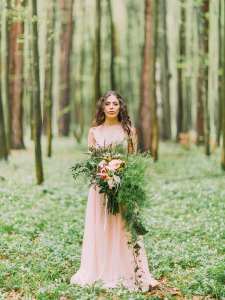 Kvinnan med långt lockigt hår i långa ljus rosa klänning håller stor bukett av röda, vita blommor och gröna element och tittar på kameran. Fullängds fotot i trä. — Stockfoto