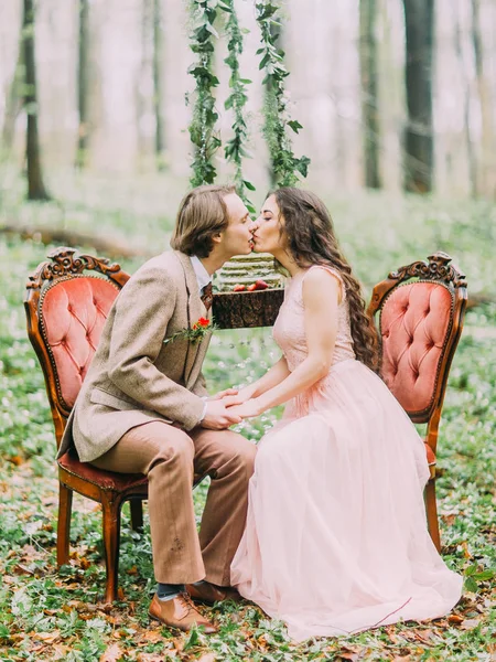 De kyssa vintage klädd nygifta sitter på de gamla snygga orange stolarna framsidan av hängande stubbe med grönt, vitt och jordgubbs kaka i gröna våren trä. — Stockfoto
