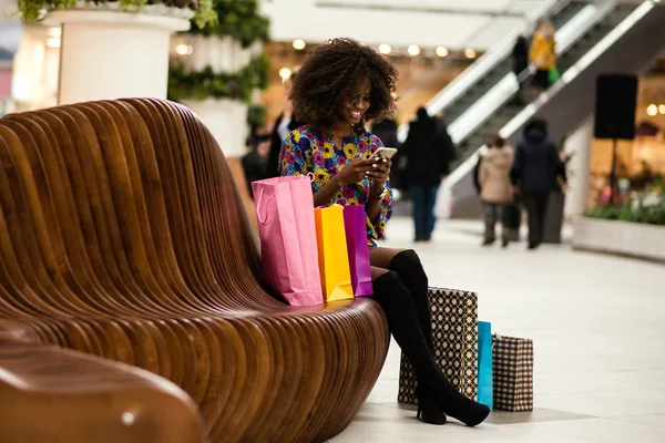 Σγουρά μαλλιά αφρο-αμερικανικό κορίτσι σε ένα εμπορικό κέντρο που κάθεται σε ένα παγκάκι με το κινητό τηλέφωνο στο herhends. — Φωτογραφία Αρχείου