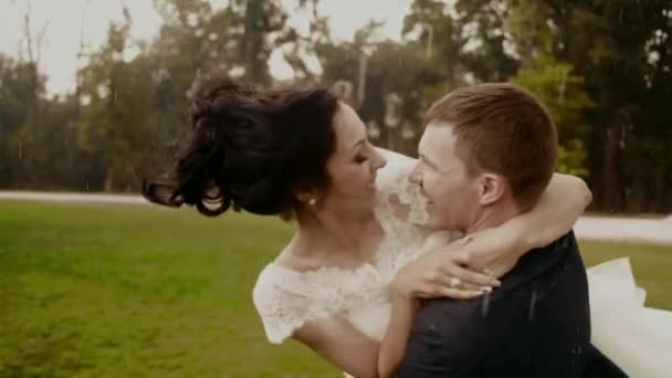 El novio feliz está haciendo girar a la novia sonriente en la lluvia en el parque durante el verano . — Vídeos de Stock