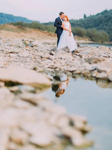 Ελκυστική νεαρή γαμήλιο ζεύγος στοργικά φαίνονται σε κάθε άλλο. Όχθη του ποταμού στο βουνό σε φόντο — Φωτογραφία Αρχείου