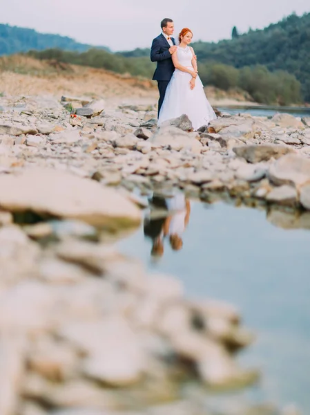 Attraente giovane coppia di nozze amorevolmente guardarsi. Riva di un fiume di montagna con pietre sullo sfondo — Foto Stock