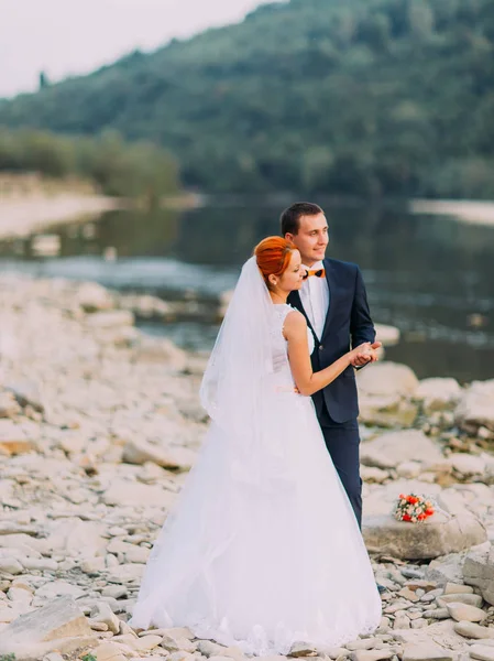 Hermosa pareja de boda abraza suavemente cerca de la orilla de un río de montaña con piedras —  Fotos de Stock