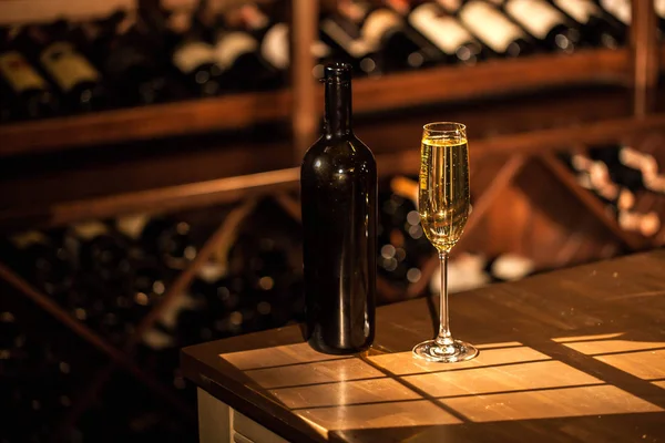 Bottiglia di champagne e bicchiere disposti su un tavolo di legno. Scaffali con bottiglie di vino su sfondo . — Foto Stock