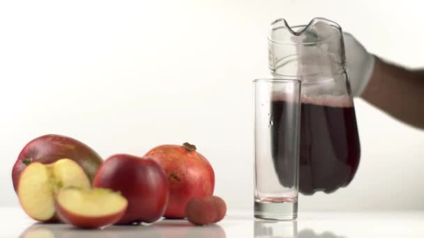 The fruit composition consisted of apples and pomegranate placed near the empty glass. The man is pouring the red juice into the glass. — Stock Video