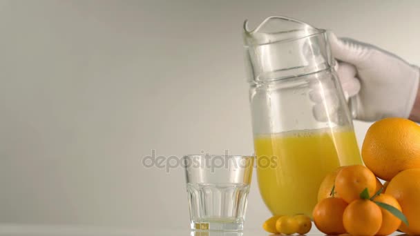 La mano en guantes blancos está vertiendo el jugo de naranja en el vaso de rocas colocado junto a la composición de la fruta consistió en las naranjas y mandarinas . — Vídeo de stock