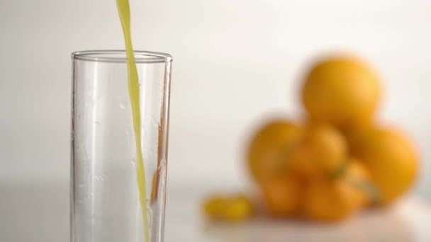 La vue rapprochée de la boisson à l'orange coulant dans le verre haut avec la grande vitesse à l'arrière-plan flou des oranges et des mandarines . — Video