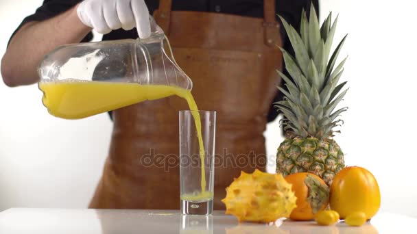 The man is pouring the orange juice from the decanter into the high glass placed near pineapple, persimmons and kiwano. — Stock Video