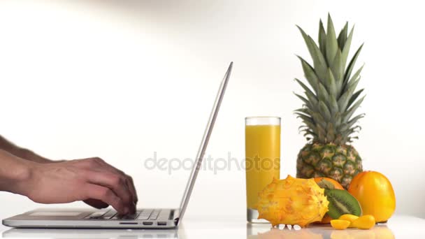 The close-up side view of the man typing on the laptop placed next to the pineapple, persimmons, kiwano and orange juice in the studio.