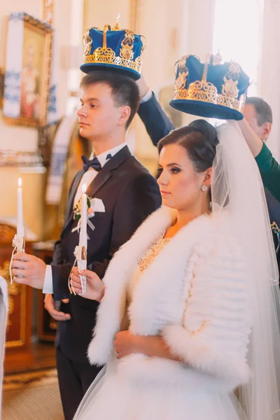Os recém-casados felizes estão segurando as velas com fitas brancas enquanto estão sob as coroas de ouro . — Fotografia de Stock