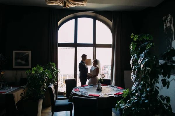 Красивые, романтичные молодожены позируют возле яркого окна в роскошном свадебном зале перед торжеством — стоковое фото