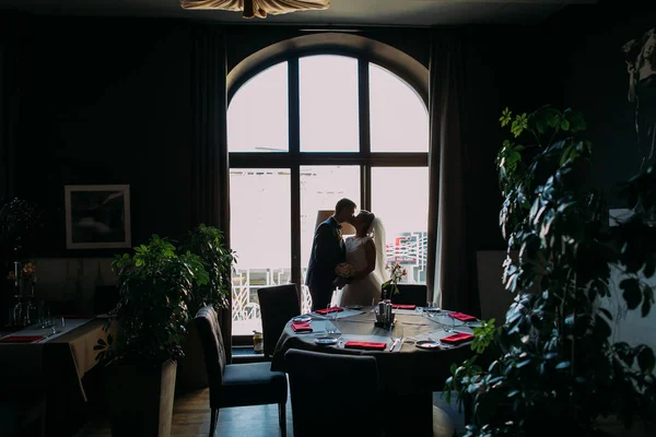Prachtige, romantische jonggehuwden kussen voor heldere raam met Bruiloftszaal voor de viering als achtergrond — Stockfoto