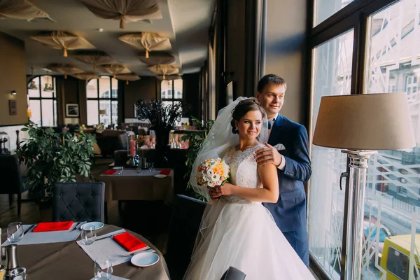 Hermosos y románticos recién casados posando cerca de la ventana en el lujoso salón de bodas antes de la celebración —  Fotos de Stock