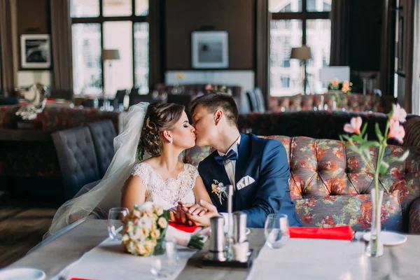 Casamento recém-casado casal apaixonado beijando enquanto sentado no sofá. Interior brilhante de luxo com grandes janelas como fundo — Fotografia de Stock