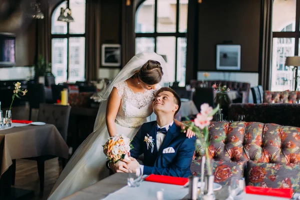 Una pareja de enamorados recién casados. Hermosa joven esposa en vestido blanco mirando a su marido suite azul posando en el interior, interior brillante de lujo con grandes ventanas —  Fotos de Stock