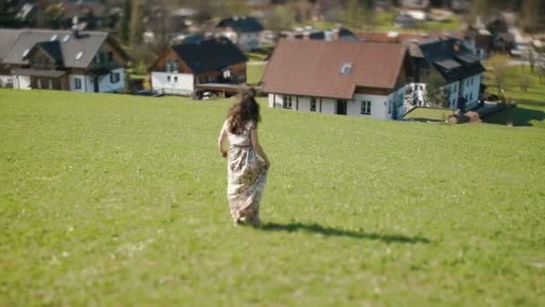 La vue arrière de la jeune fille dans la longue robe rustique tournant autour de la prairie à l'arrière-plan des montagnes couvertes de maisons . — Video