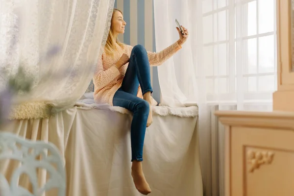 The young teenager is sitting on the bed and taking the selfie via the phone. — Stock Photo, Image