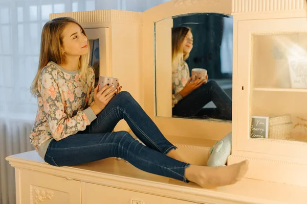 Горизонтальна фотографія мрійливої молодої дівчини, що тримає чашку чаю і сидить на туалетному столику . — стокове фото