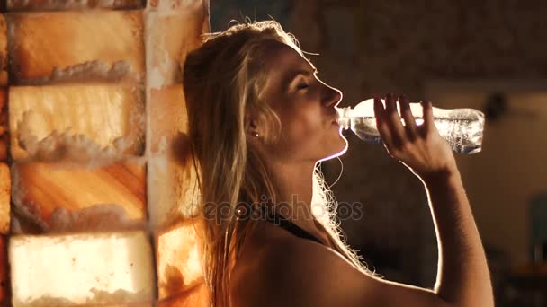 The side portrait of the woman drinking water from a bottle and leaning on the wall in the dark room. — Stock Video