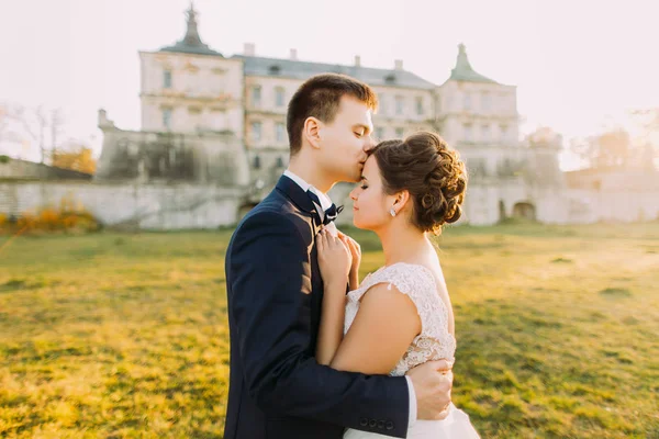 O noivo está beijando a bela noiva na testa no fundo do antigo castelo gótico. durante o pôr-do-sol. Vista de perto — Fotografia de Stock