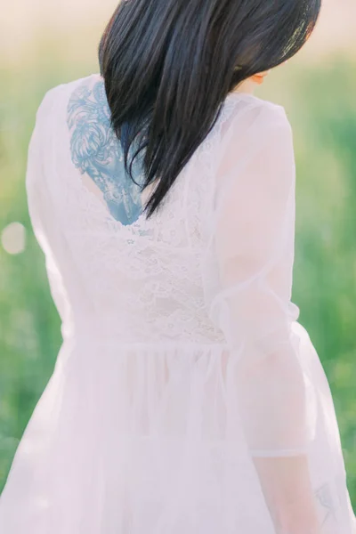 Die Nahaufnahme der Braut im weißen Kleid mit den offenen Schultern und zurück im grünen Frühlingsfeld. das Tattoo auf dem Rücken. — Stockfoto