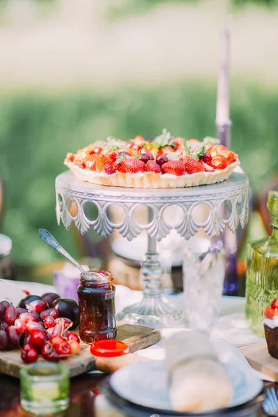 Der herrliche Biskuitkuchen aus Erdbeeren und Kirschen steht auf dem weißen Kuchenständer vor dem verschwommenen Hintergrund des grünen Waldes. — Stockfoto