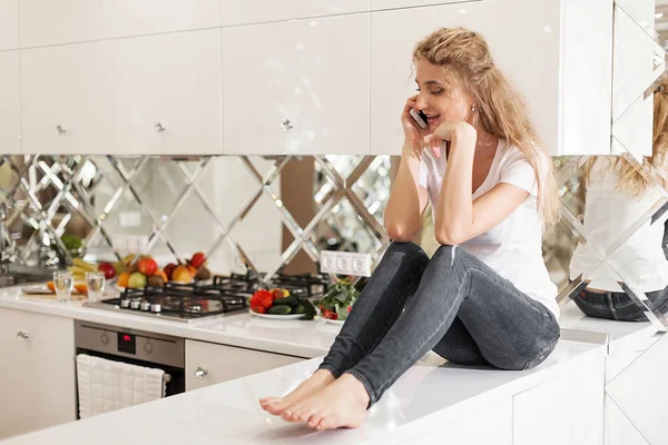 Genç sarışın mutfak yüzeyinde oturan ve telefonda konuşurken gülümsedi. Beyaz T-shirt ve jeans giymiş kadın. — Stok fotoğraf