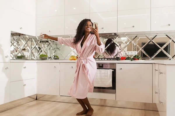 Happy young black woman wearing bathrobe dansing in the kitchen with the phone in her hands. — Stock Photo, Image