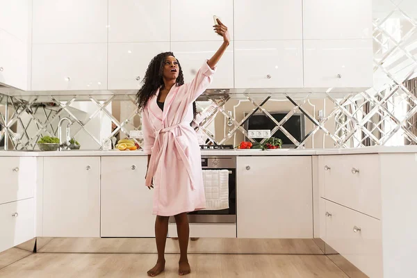 Attractive young black woman with long curly hair standing in the kitchen and making selfie. Woman wearing pink bathrobe. — Stock Photo, Image