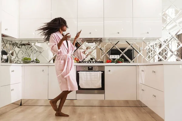 Slender black woman dancing in the kitchen with the telephone and glass of water in her hands. — Stock Photo, Image